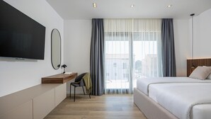 Un dormitorio de hotel moderno con una cama grande, un escritorio con una silla, un televisor montado en la pared y un ventanal con cortinas.