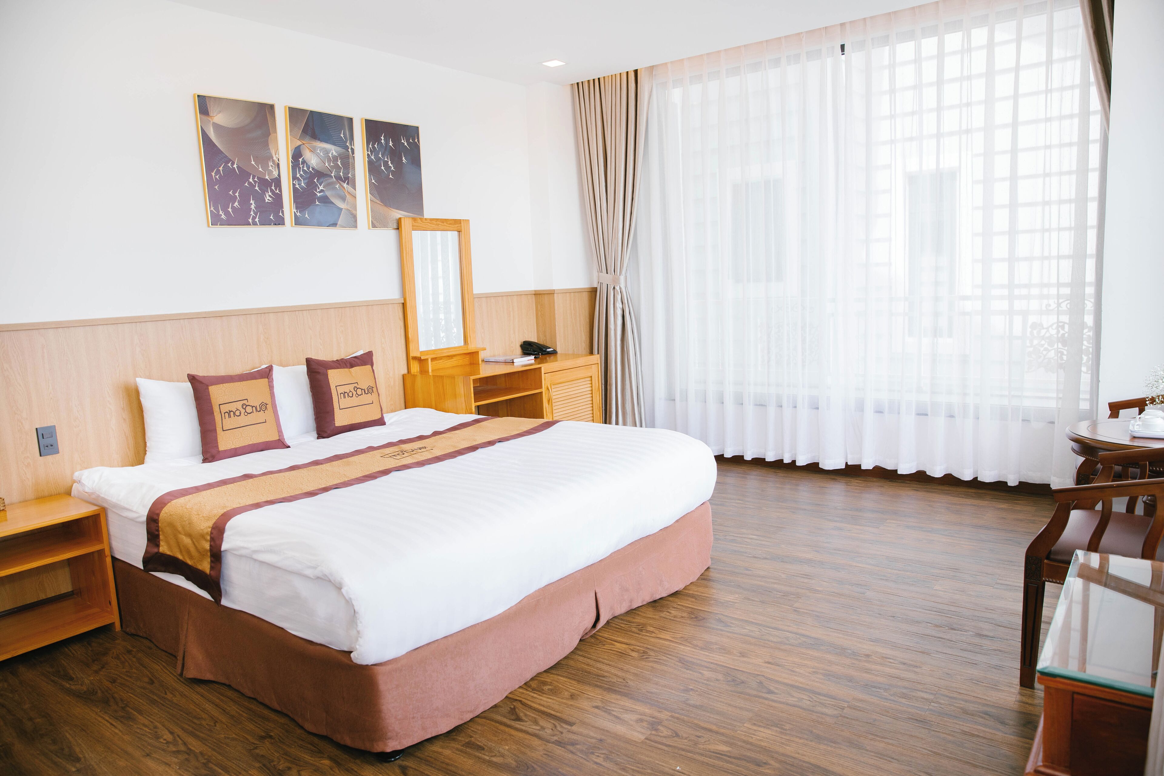 A hotel room with a large bed, wooden headboard, a bedside table, a nightstand, a desk with a chair, and a window with sheer curtains.