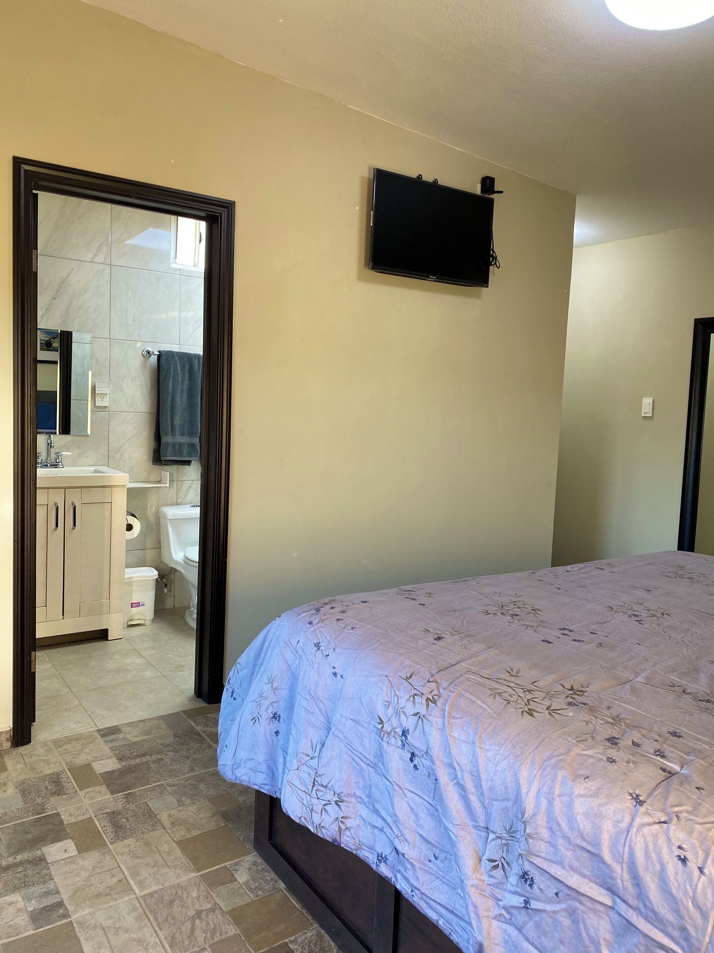 Een slaapkamer met een bed, een televisie aan de muur, een badkamer met toilet, wastafel en spiegel, en een deur die naar een andere kamer leidt.