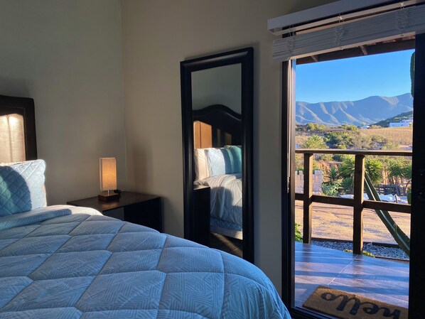 Un dormitorio con cama, mesita de noche, espejo y vistas a las montañas a través de un ventanal abierto.