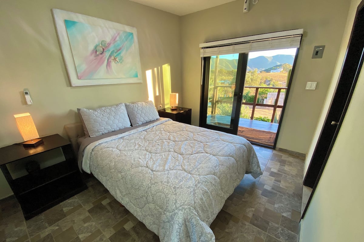A bedroom with a large bed, a nightstand, a painting on the wall, and a view through a sliding glass door.