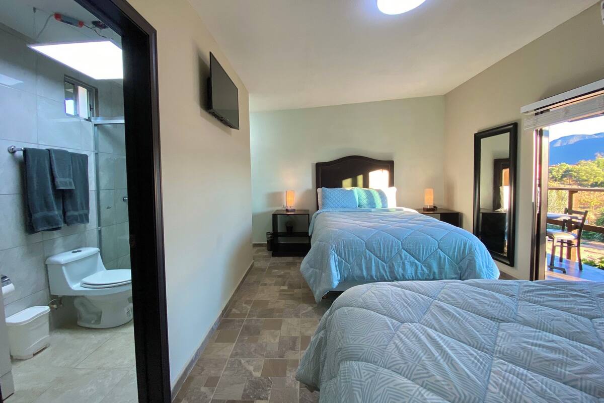 A bedroom with a bed, a bedside table with a lamp, a TV mounted on the wall, and a bathroom with a toilet and towel rack.