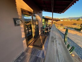 A covered outdoor seating area with a wooden deck, a glass door entrance, and a view of the surrounding landscape.