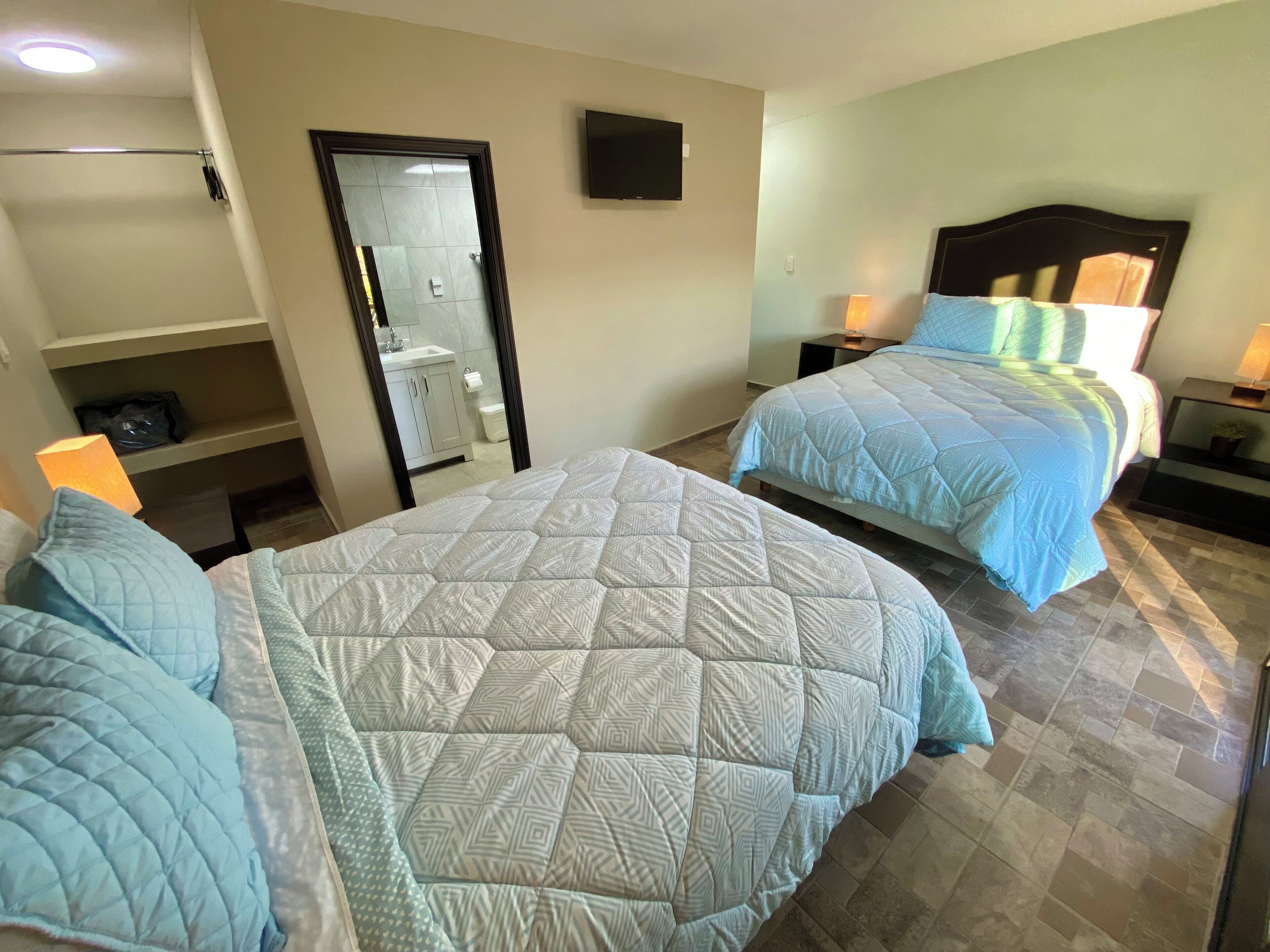 Una camera d'albergo con due letti, un bagno, una TV e uno scaffale con una borsa.