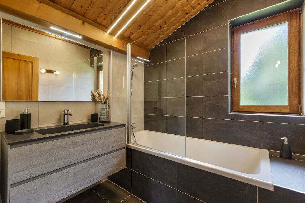 A modern bathroom with a wooden ceiling, a large mirror, a double sink vanity, a bathtub, and dark tiled walls.