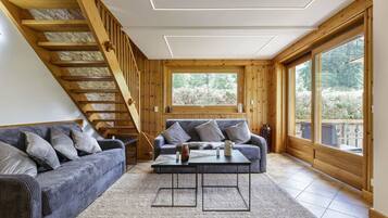 A modern living room with a wooden staircase, a grey sofa, a coffee table, and large windows offering a view of the outdoors.