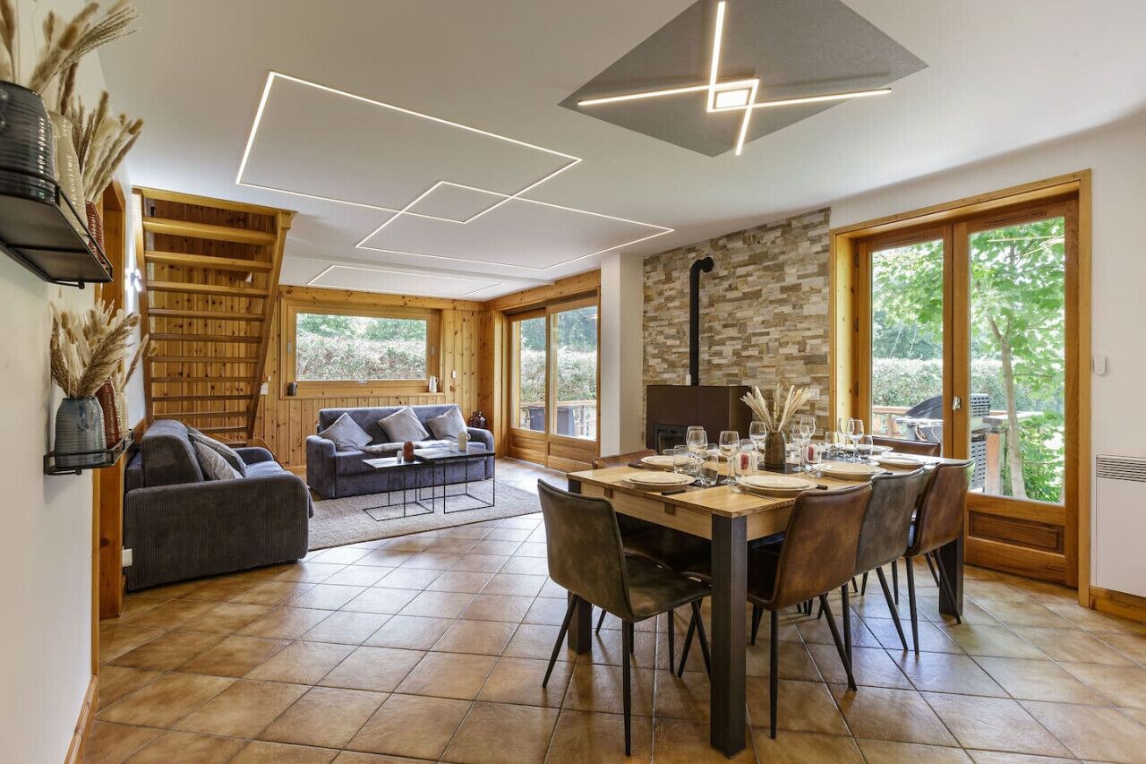 A modern dining area with a wooden table set for four, surrounded by chairs. There is a sofa and a small table with a lamp in the background.