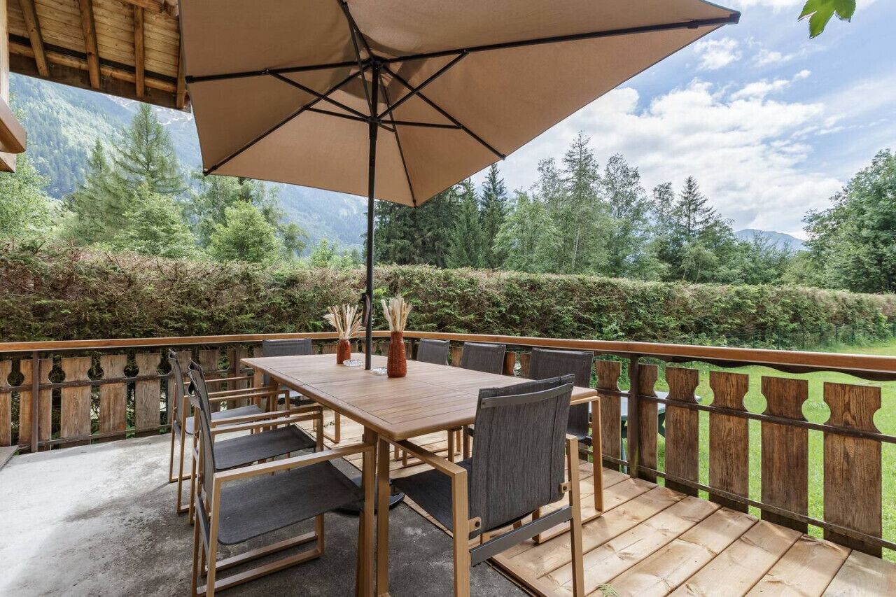A wooden deck with a table and chairs, a large umbrella, and a view of a forested area with mountains in the background.
