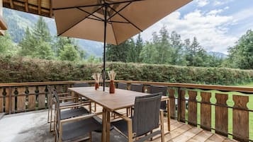 A wooden deck with a table and chairs, a large umbrella, and a view of a forested area with mountains in the background.