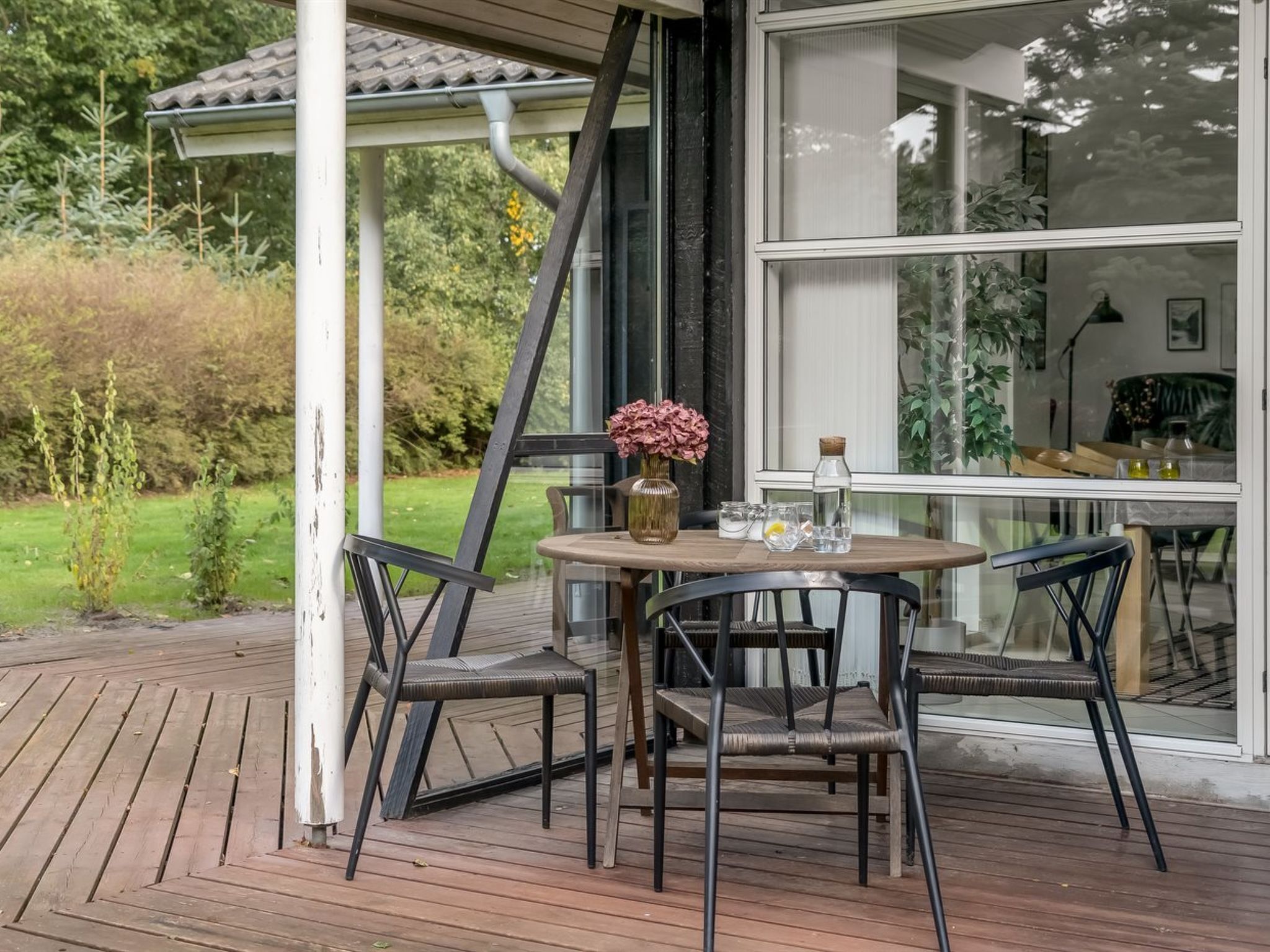 Une terrasse en bois avec une table et des chaises, une porte vitrée donnant sur une pièce et un jardin en arrière-plan.