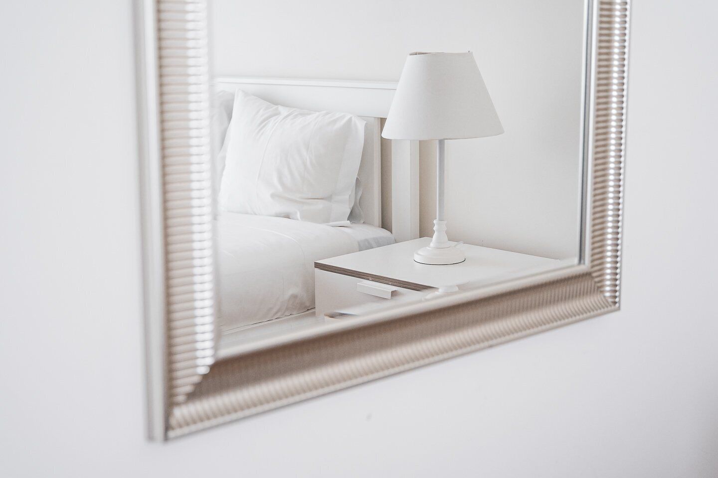 Une table de chevet blanche, sur laquelle se trouvent une lampe et un miroir qui reflète un lit recouvert de linge de lit blanc.