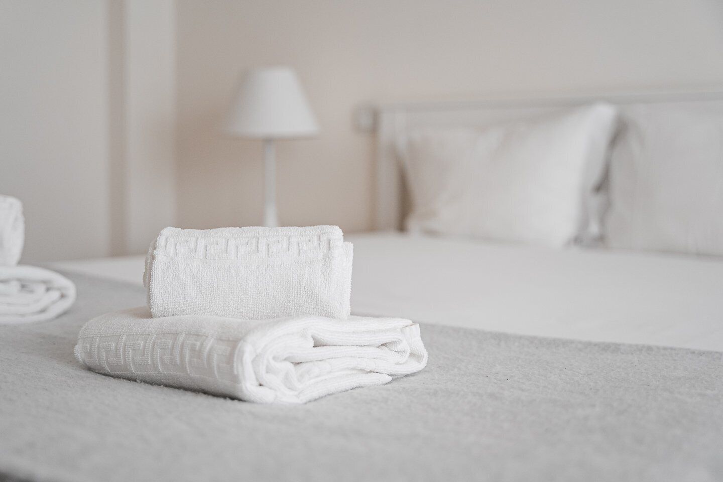 Una cama bien tendida con sábanas blancas, una lámpara blanca y un almohadón sobre una colcha blanca.