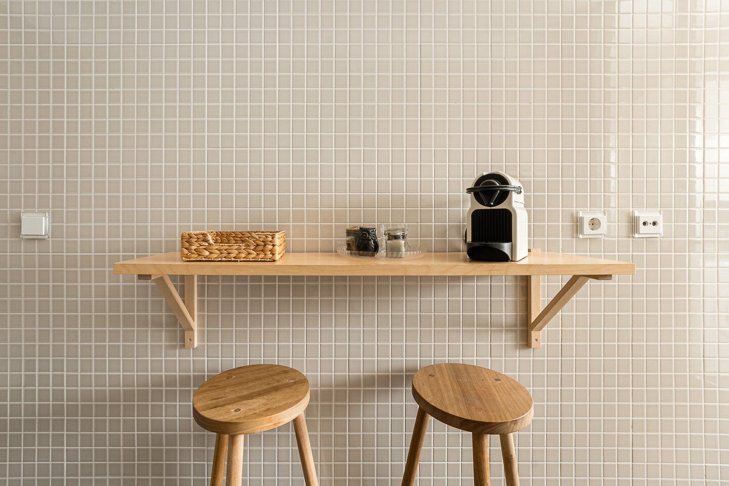 Une cuisine avec une étagère en bois sur laquelle sont posés un panier tressé, une machine à café et un bocal en verre. On peut voir deux tabourets en bois devant.