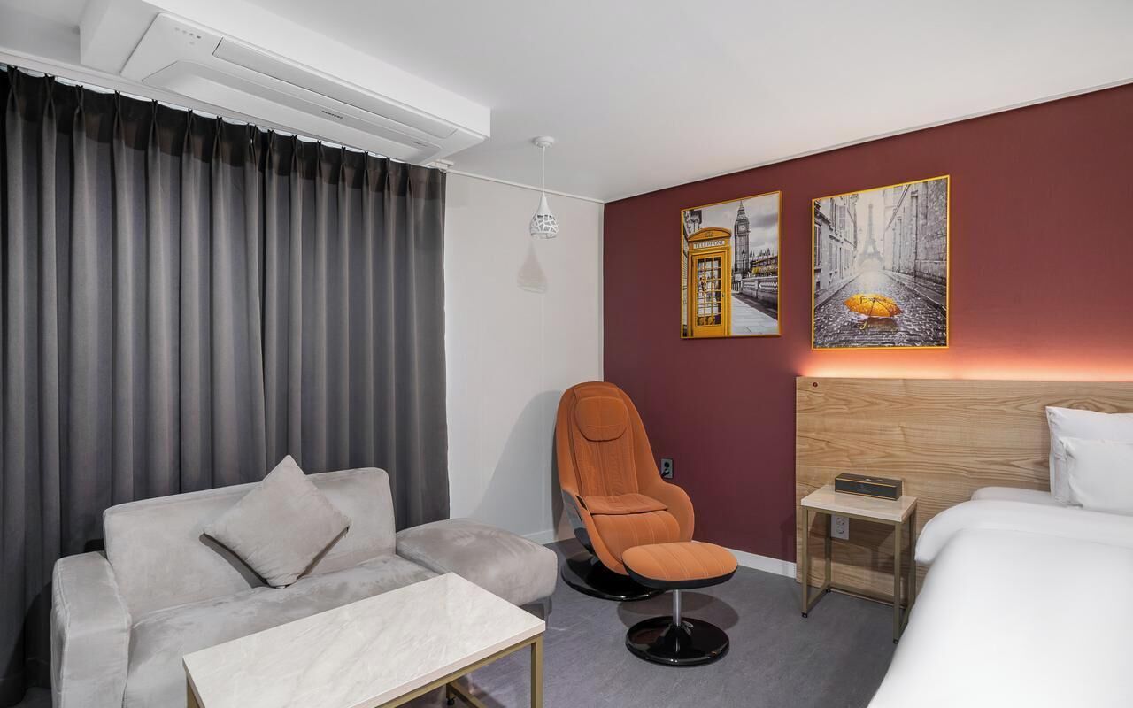 A hotel room with a sofa, armchair, bedside table, and a bed. There are two paintings on the wall and a ceiling light.