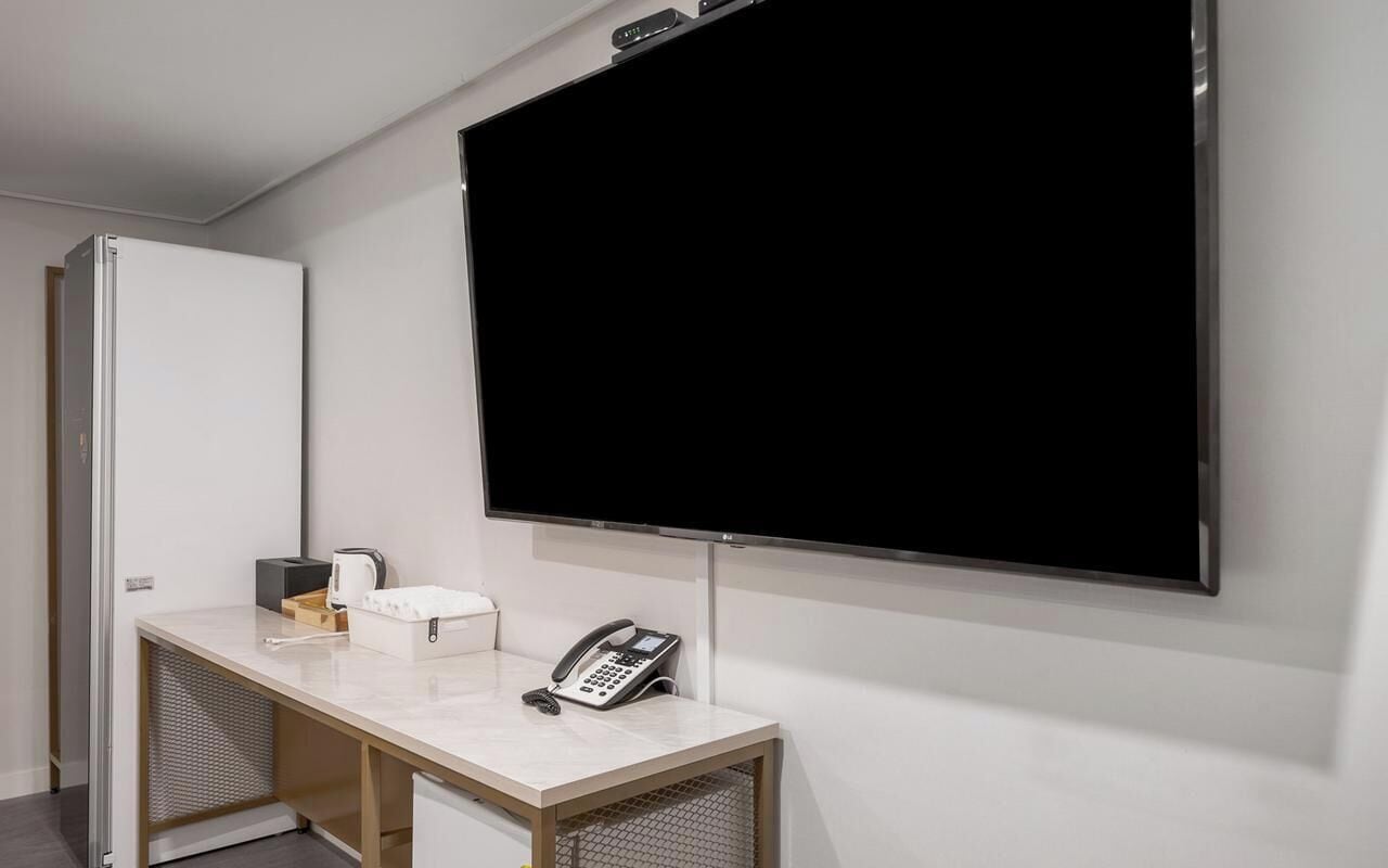 Et hotelværelse med et stort fladskærms-tv på væggen, et skrivebord med telefon, kaffemaskine og en lille æske.