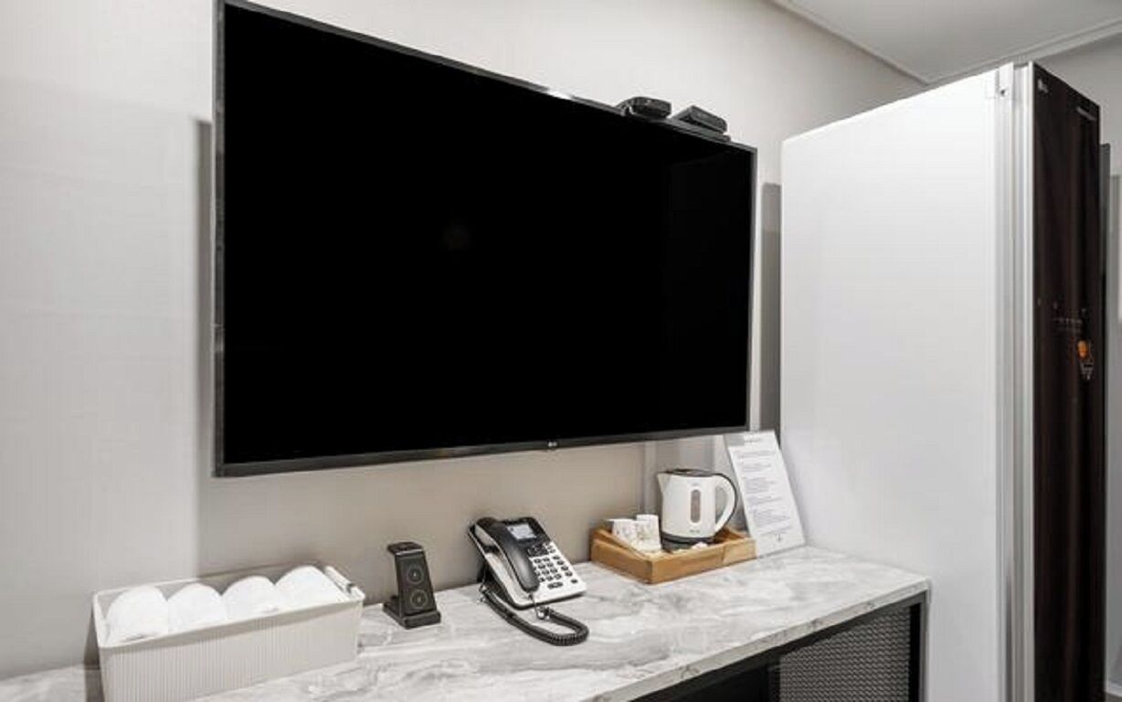 Habitación de hotel con televisor de pantalla plana grande montado en la pared, teléfono, cafetera y bandeja con tazas y un menú sobre la encimera de mármol.