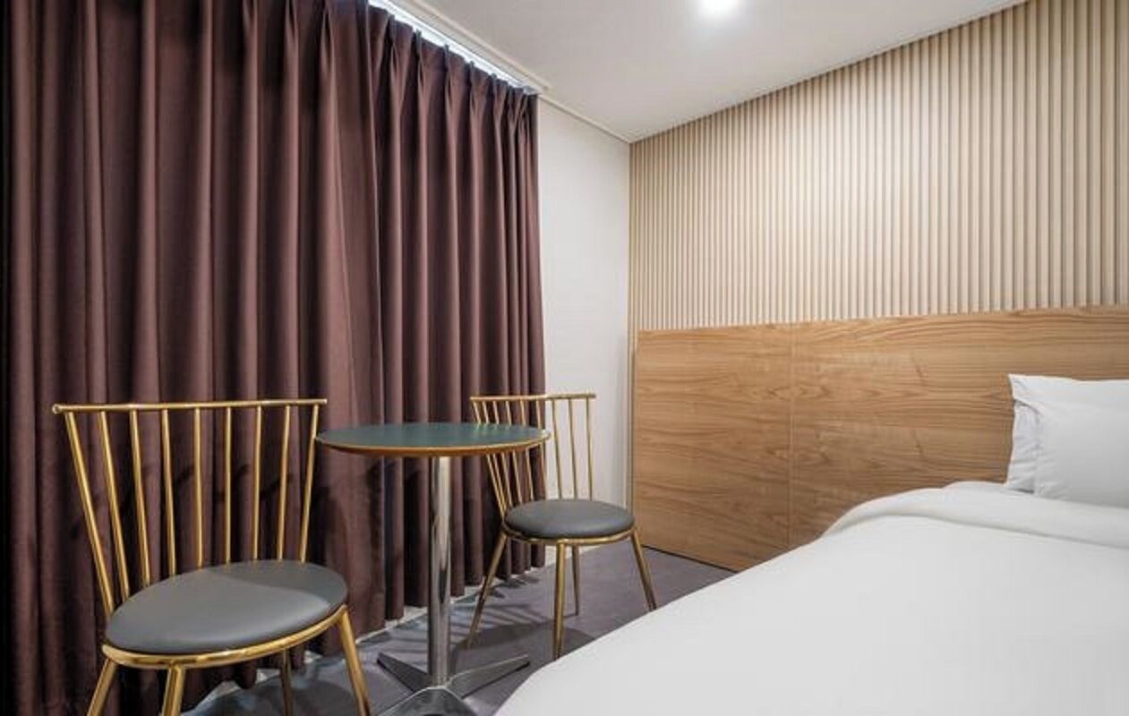 Una habitación de hotel con una cama, dos sillas, una mesita y una cortina.