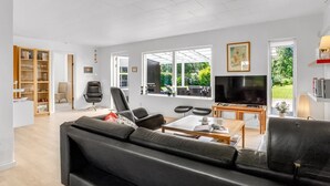 Un salon spacieux avec un canapé en cuir noir, une table basse en bois et un téléviseur. On aperçoit une bibliothèque et un fauteuil au fond de la pièce.