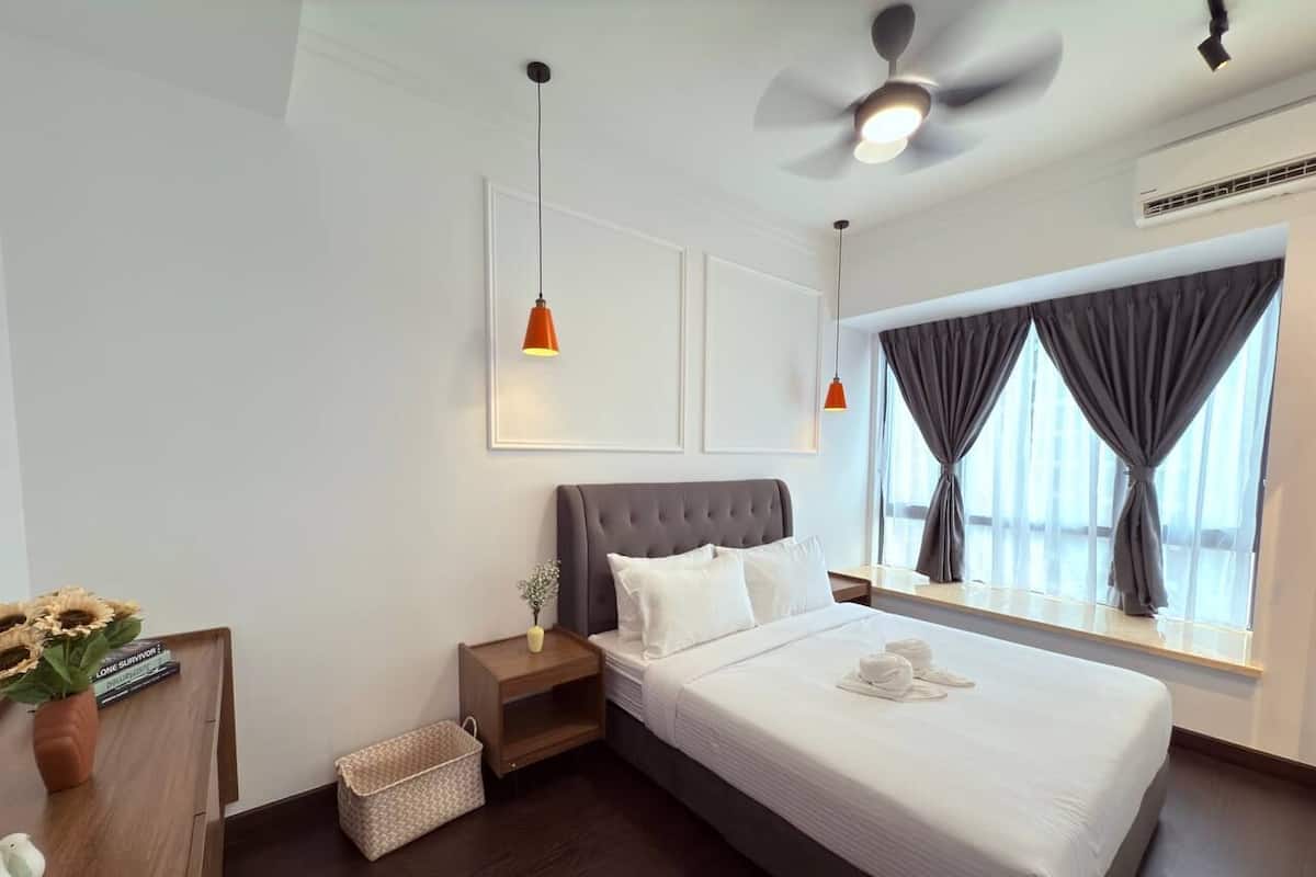 A hotel room with a large bed, a ceiling fan, a window with curtains, a bedside table with a lamp, and a small plant.