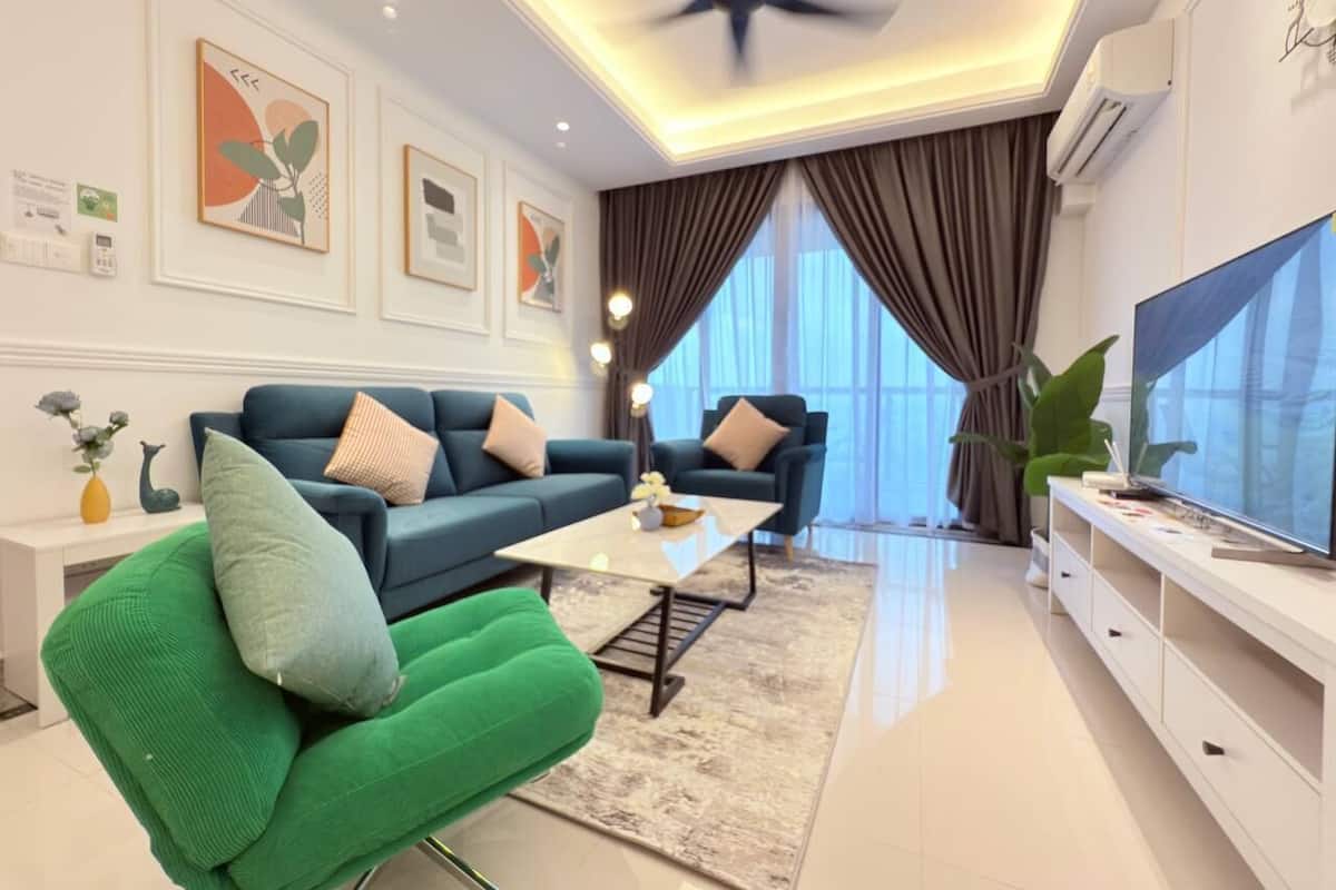 A modern living room with a blue sofa, green armchair, and a white coffee table. There is a flat-screen TV on a white stand, and the room has large windows with curtains.
