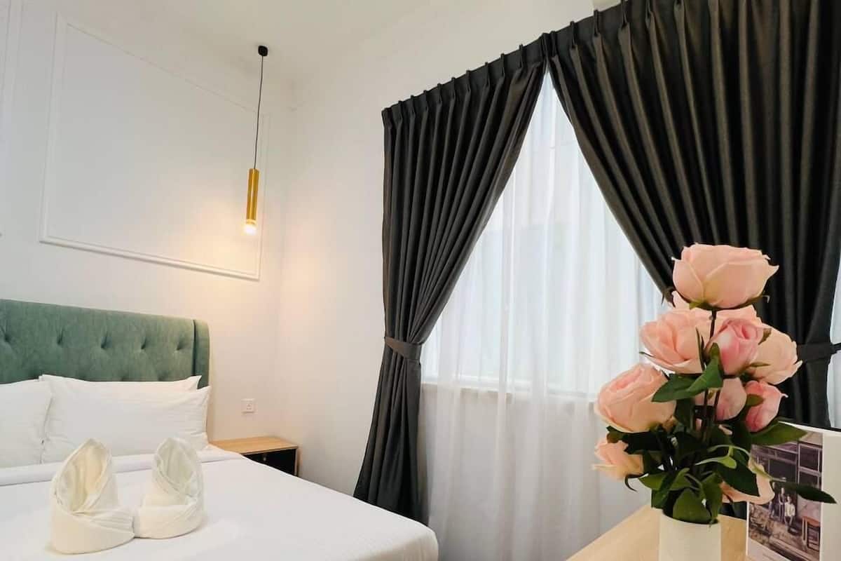 A hotel room with a bed, a bedside table with a vase of flowers, and a window with curtains.