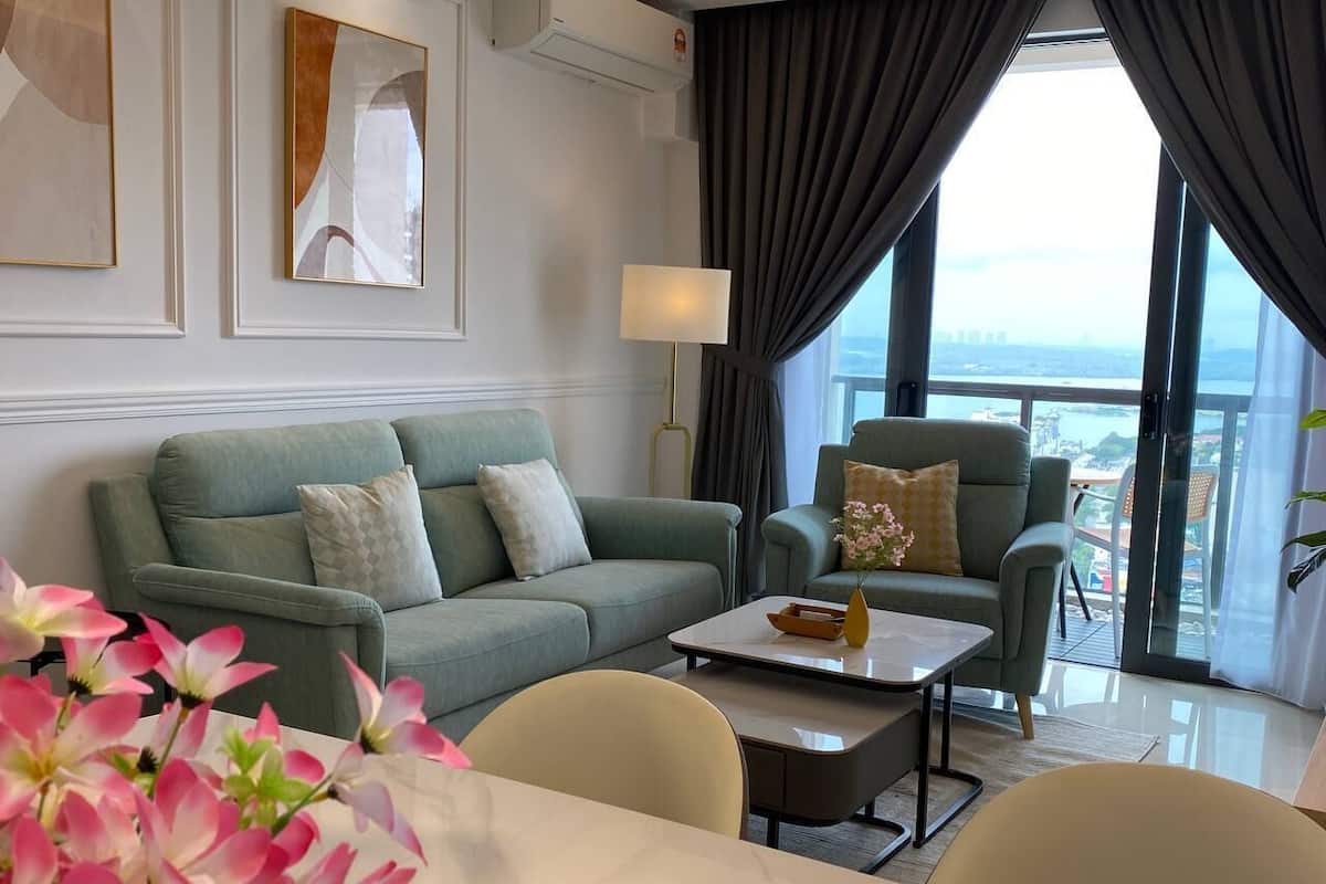 A modern hotel room with a green sofa, a matching armchair, a dining table, and a view of a cityscape through large windows.
