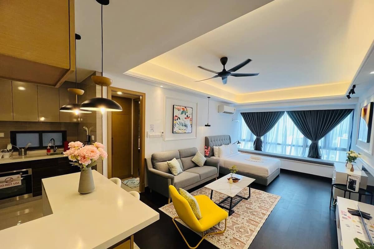 A modern living room with a sofa, coffee table, and yellow chair. There is a dining area with a white table and a vase of flowers. The room has a ceiling fan and large windows with curtains.