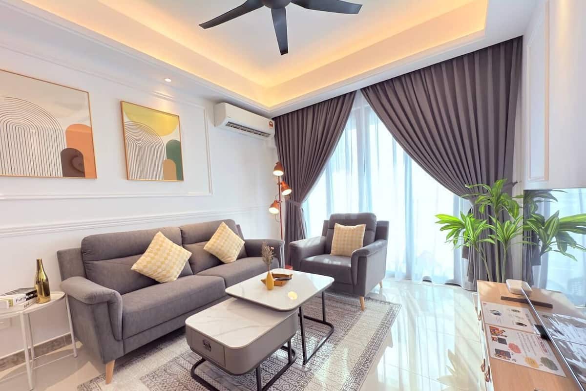 A modern living room with a grey sofa, a white coffee table, and a grey armchair. There is a small side table with a bottle and a magazine. The room has a ceiling fan, a large window with curtains, and a potted plant.