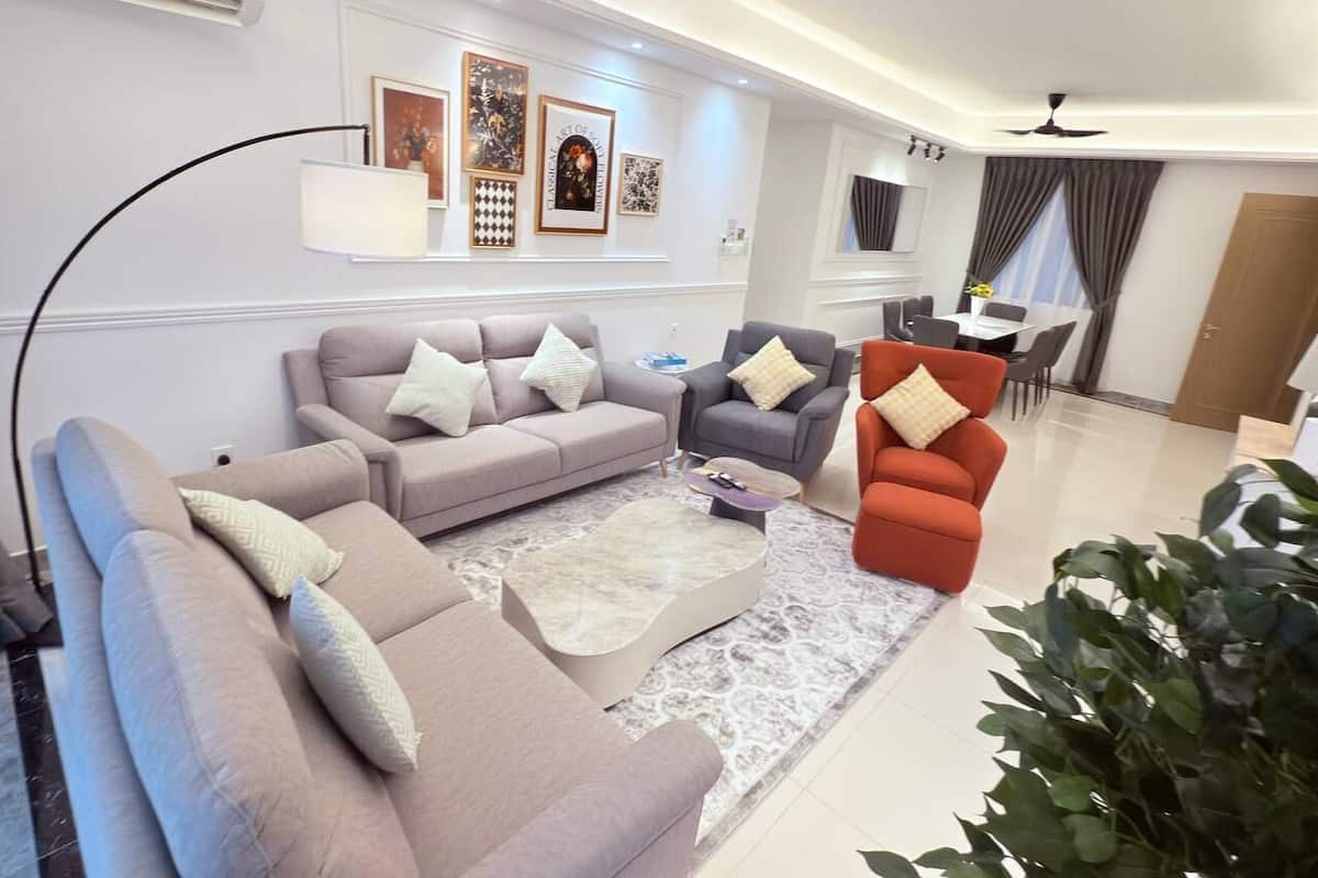 A modern living room with a grey sofa, a red armchair, and a coffee table. There are framed artworks on the wall and a ceiling fan.