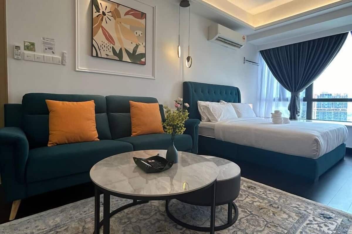 A modern hotel room with a teal sofa, a round coffee table, a bed with a blue headboard, and a large window with curtains.