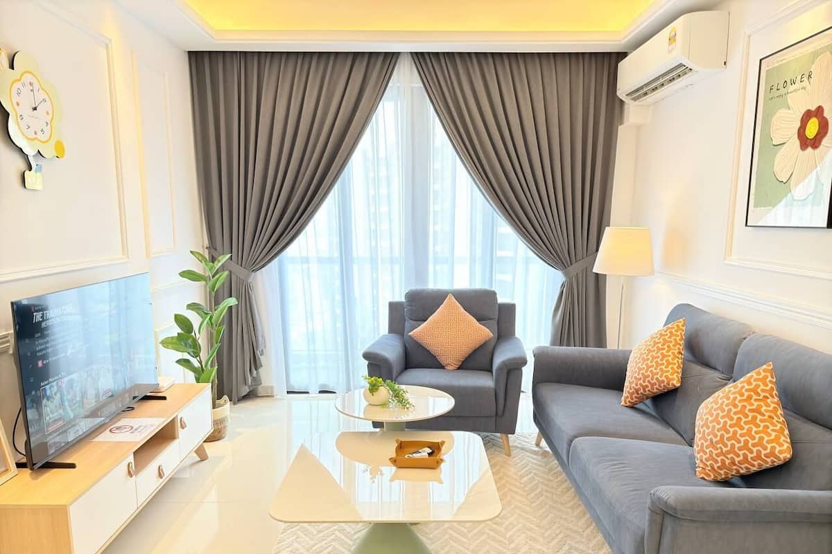 A modern living room with a flat-screen TV, grey sofas with patterned cushions, a coffee table, and a large window with grey curtains.