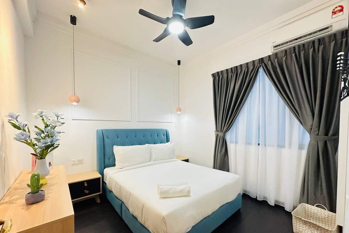 A hotel room with a single bed, a ceiling fan, a bedside table with a lamp, a window with curtains, and a small plant on the nightstand.