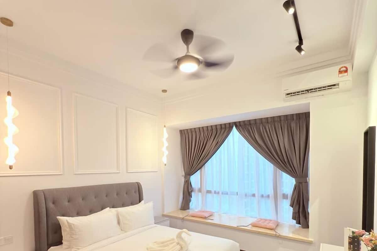 A modern hotel room with a large bed, a ceiling fan, a window with curtains, a bedside table, and a small desk with a lamp.