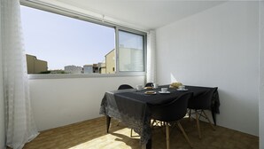 A dining area with a table set for two, a window with a view of buildings, and a curtain on the left.