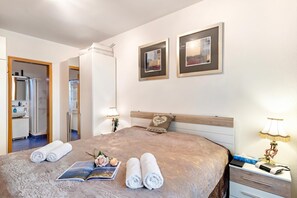 A hotel room with a bed, a nightstand, a lamp, a framed picture on the wall, and a bathroom visible through an open door.