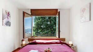A bedroom with a large window, a bed with a red quilt, a bedside table with a lamp, and a framed artwork on the wall. - Vernate Lugano Lake Sweet View (Vernate)
