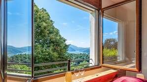 A room with a large window overlooking a mountainous landscape, a wooden bench, and a bed with a pink patterned bedspread. - Vernate Lugano Lake Sweet View (Vernate)