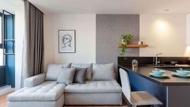 Un salon moderne avec un canapé d’angle gris, un petit îlot central doté d’un évier et une œuvre d’art encadrée accrochée au mur.