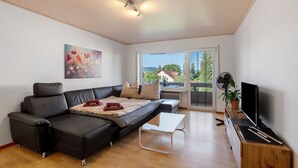 Een moderne woonkamer met een bank, een flatscreen televisie en een raam met uitzicht op een huis en bomen.