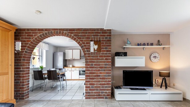 A living room with a TV, white entertainment unit, and a brick archway leading to a kitchen.