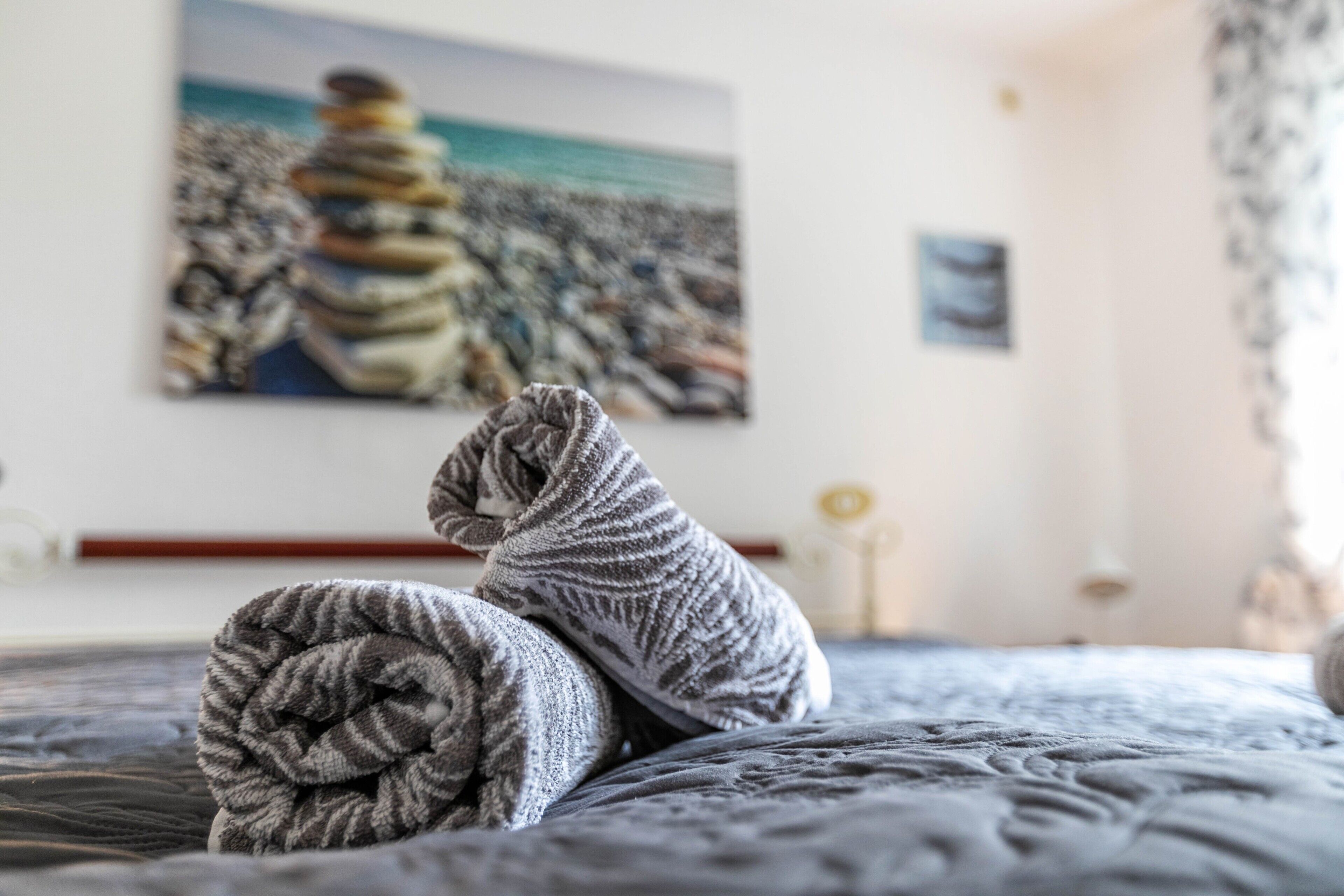 A bed with rolled towels and a painting of stacked stones on the wall.