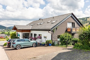 Een tweelaags huis met een schuin dak, een balkon op de tweede verdieping en een oprit met twee geparkeerde auto's.
