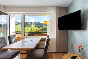 A modern room with a dining table, chairs, a flat-screen TV, and a large window offering a view of a green landscape.