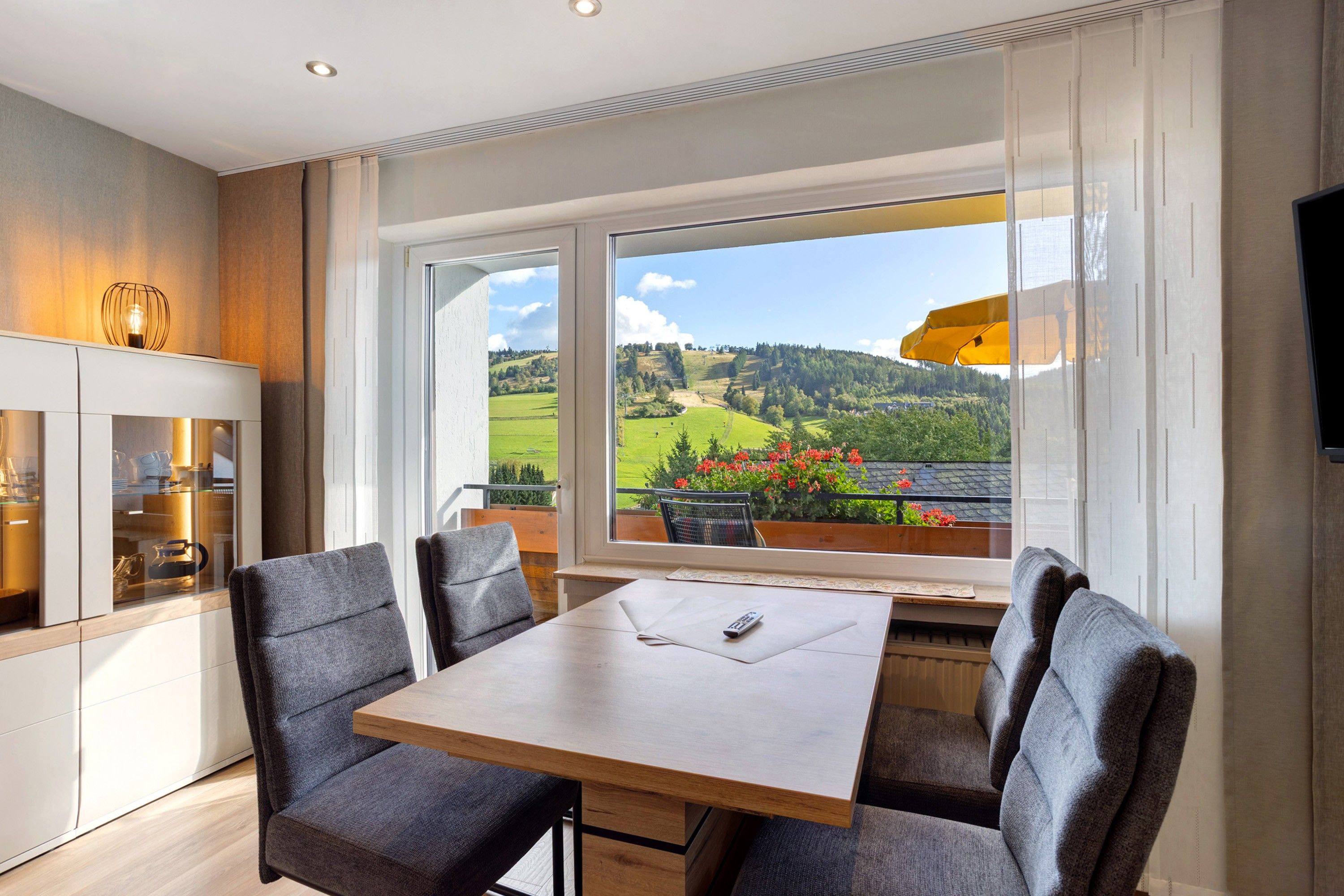 Ein Essbereich mit Tisch und Stühlen, Blick auf eine grüne Landschaft durch große Fenster sowie eine Küchenzeile mit Schränken und einer Hängeleuchte.