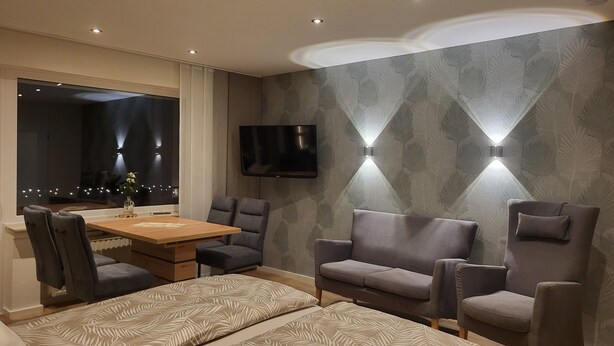 A modern hotel room with a wooden dining table, two gray armchairs, a sofa, a TV mounted on the wall, and a patterned wallpaper. - Haus Schönblick – Apartment 3 with Mountain View, Balcony and Wi-Fi (Willingen (Upland))