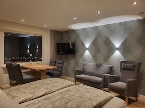 Een moderne hotelkamer met een houten eettafel, twee grijze fauteuils, een bank, een aan de muur bevestigde televisie en behang met een patroon.