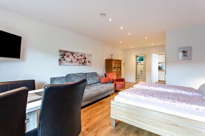 A modern living room with a sofa, a dining table with chairs, a flat-screen TV mounted on the wall, and a bed with a floral bedspread.