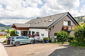 Een tweelaags huis met een schuin dak, een balkon op de tweede verdieping en een oprit met twee geparkeerde auto's.