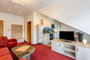 Een ruimtelijk ingerichte leefruimte met een kitchenette, een flatscreen televisie, een bank en een kamerbreed tapijt met dessin.