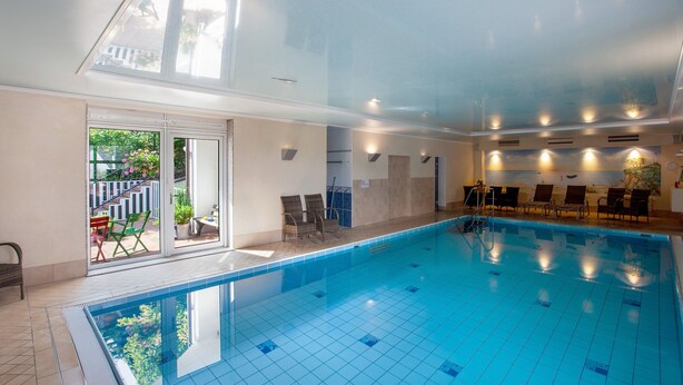 Ein Hallenbad mit großen Glaswänden, gefliestem Boden und einer Sitzecke mit Stühlen.